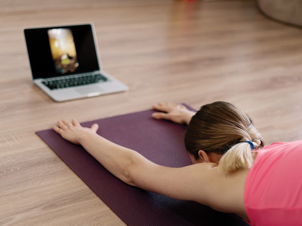 Woman doing yoga child's pose in front of laptop set on the floor. 