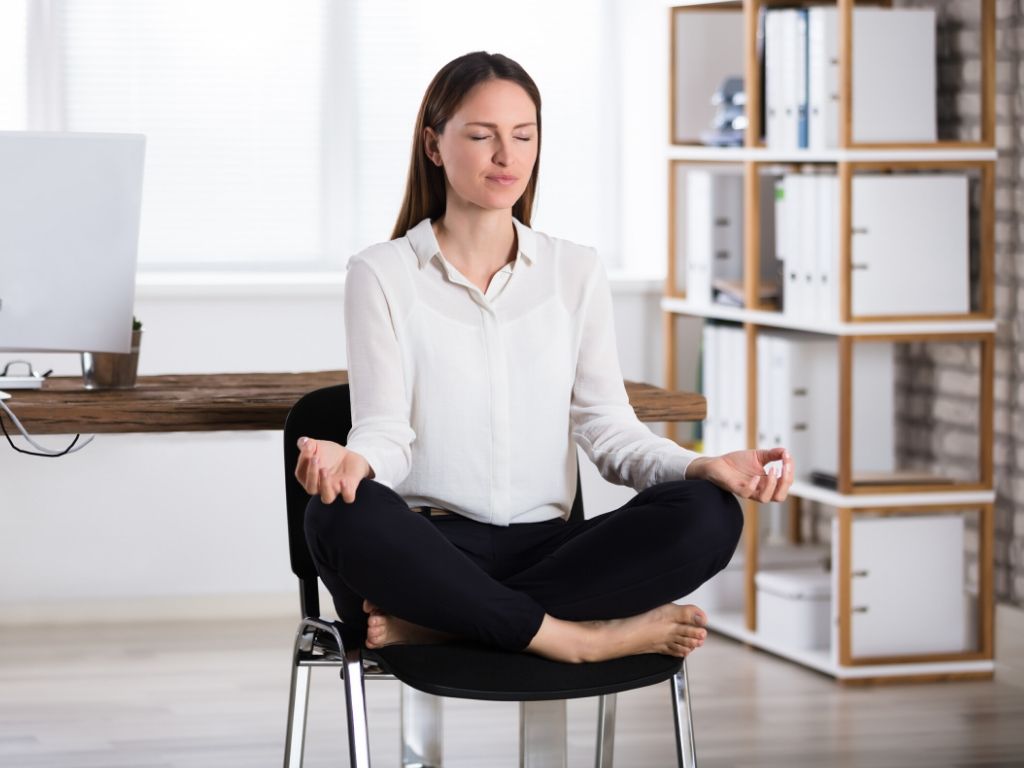 Business woman doing cross legged meditation yoga pose seated on a chair. 