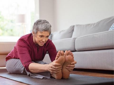 Woman doing yoga stretch, sitting on the floor bent at waist touching feet, in home living room in front of couch on a yoga mat.